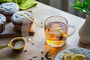 A cup of tea with lemon and cupcakes on a table near the window.