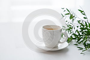 A cup of tea with lemon and green branch on a white table against the background of a kitchen window