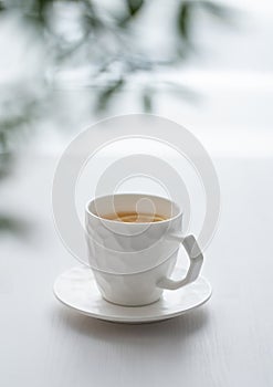 A cup of tea with lemon and green branch on a white table against the background of a kitchen window