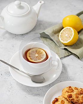 A cup of tea with lemon and homemade cookies on a light background with a white teapot and citrus close up. Healthy morning drink