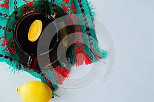 A cup of tea with lemon stands on a dark saucer, which stands on a knitted green-red scarf, next to it lies a lemon on a