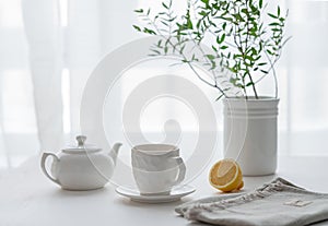 A cup of tea with lemon, a teapot and green branch on a white table against the background of a kitchen window