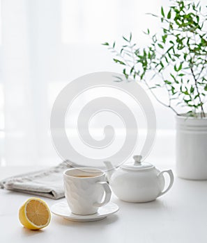 A cup of tea with lemon, a teapot and green branch on a white table against the background of a kitchen window