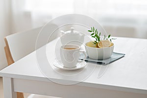 A cup of tea with lemon and a teapot on a white table against the background of a kitchen window