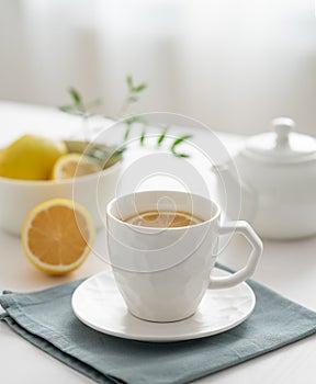 A cup of tea with lemon and a teapot on a white table against the background of a kitchen window close up