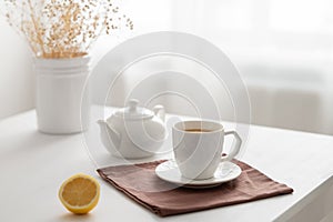 A cup of tea with lemon and a teapot on a white table with dry bouquet against the background of a kitchen window