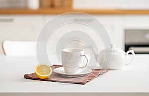 A cup of tea with lemon and a teapot on a white table. Kitchen with wooden empty countertop and brick wall in the background