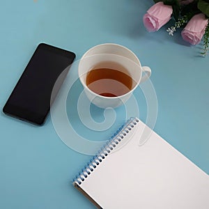Cup of tea, smartphone, blank notepad, roses on blue background.