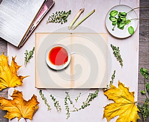 Cup of tea standing on an open book with autumn leaves, books, mint and thyme on wooden rustic background top view