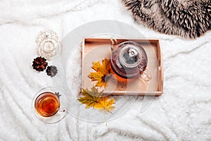 Cup of tea and teapot in wooden tray on the bed. Warm sweater, candles, yellow leaves. The concept of cozy autumn weekend house.