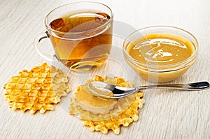 Cup with tea, waffle, bowl with peanut butter, spoon with peanut paste on waffle on wooden table