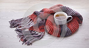 A cup of tea wrapped in a wool terracotta color scarf stands on a beige wooden surface. View from above