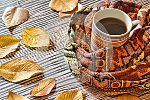 cup of warm coffee wrapped in a scarf on a wooden table with fallen autumn leaves