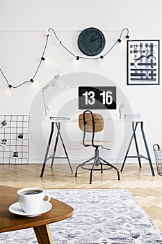 Cup on wooden table in white home office interior with chair at desk with desktop computer. Real photo