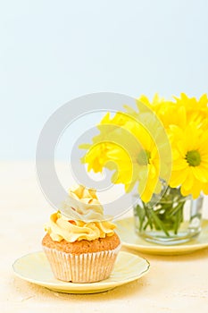 Cupcake with yellow cream decoration and bouquet of yellow chrysanthemum in glass on tender pastel background.