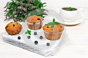 Cupcakes with black currants on a white wooden cutting board against the background of a cup and mint flowers.