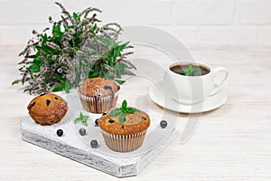 Cupcakes with black currants on a white wooden cutting board against the background of a cup and mint flowers.