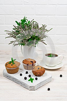 Cupcakes with black currants on a white wooden cutting board against the background of a cup and mint flowers.