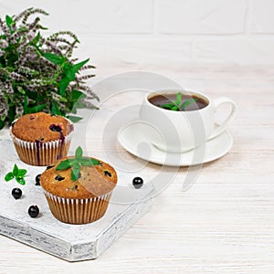 Cupcakes with black currants on a white wooden cutting board against the background of a cup and mint flowers.