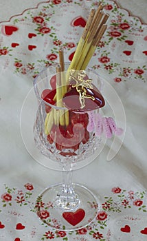 Cupid stands on enamel red heart. Candy and cookie sticks are in the wine glass. Background is all hearts on white.