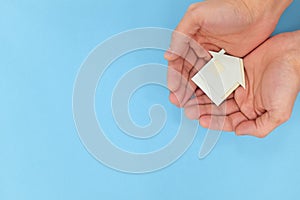 Cupped hands holding a house cutout model in blue background. Top view with copy space.