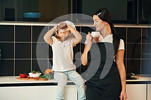 With cups, drinking. Mother with her daughter are preparing food on the kitchen