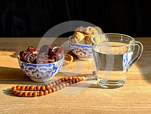 Cups with a traditional oriental pattern with dates and walnuts, a glass of clear water, stone rosary on the table during sunset