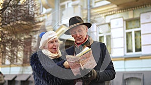 Curios couple of senior tourists standing on city street with paper map talking. Positive Caucasian elegant husband and