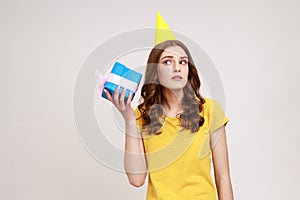 Curious adorable woman of young age in yellow casual T-shirt shaking blue giftbox near ear trying to guess her present,