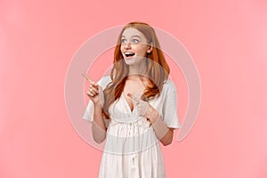 Curious and amused caucasian foxy girl in white dress, pointing fingers left looking fascinated, checking out beautiful