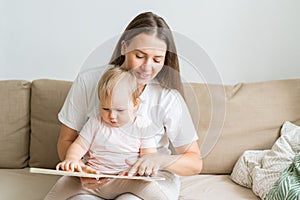 Curious baby girl sitting on mother knees and reading book with pictures. Smiling mom. Copy space