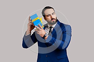 Curious bearded businessman holding and shaking wrapped present box, being interested what inside.