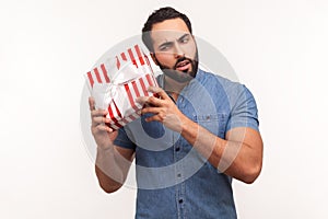 Curious bearded man shaking striped gift box trying to guess what is inside, wondered with present, surprises and bonuses