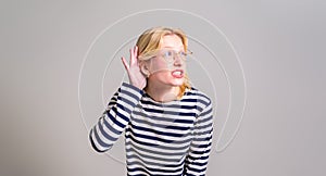 Curious blond young woman with hand near ear trying to overhead while standing on white background
