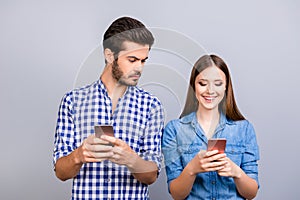 Curious boyfriend is spying his lover`s smartphone. They are wearing casual shirts, standing isolated on pure background