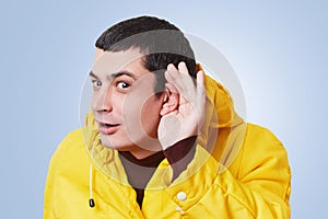 Curious brunet male model tries to overhear something, keeps hand on ear, listens gossips in next room, wears yellow anorak, isola