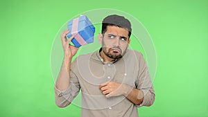 Curious brunette man with bristle in shirt shaking wrapped gift box, listening to guess what`s inside, looking inquiring confused,