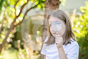 Curious clever elementary school age child, girl looking at the camera through a magnifying glass, holding a loupe in hand