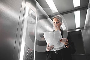 Curious corporate businesswoman standing in the elevator skeptically looking at the documents. Young female employer is critically