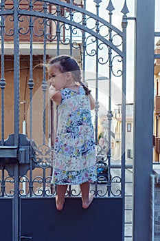 Curious, cute, barefoot blonde little girl climbing on metal fence, gate in urban city area against houses and streets