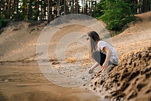 Curious girl with brown hair squatting down on sand near river in forest and going to take grit. Water reflect child.