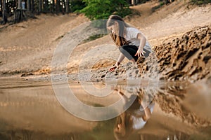 Curious girl with brown hair squatting down on sand near river in forest and going to take grit. Water reflect child.