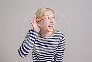 Curious happy blond young woman with hand near ear trying to listen gossip over white background