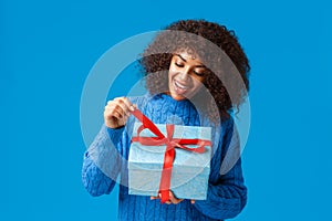 Curious and happy, smiling african-american woman, b-day girl in winter sweater, pulling present knot to unwrap gift and