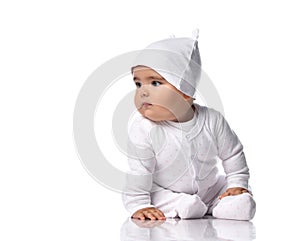 Curious infant baby toddler sits on the floor in white onepiece jumpsuit overall and funny hat with ears and looks aside