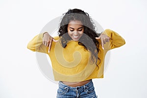 Curious and intrigued cute feminine african american girl in yellow sweater pointing and looking down with amused smile