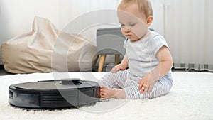 Curious little baby boy looking at robot vacuum cleaner riding on carpet at living room