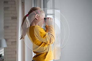Curious little girl with Down syndrome with binoculars looking through window at home.