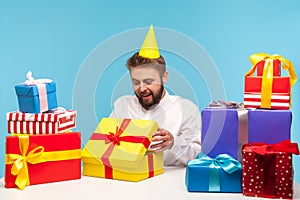 Curious man birthday boy in party hat opening gift box and looking inside, enjoying surprise celebrating holiday, receiving many