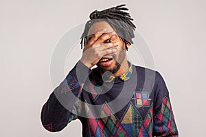 Curious nosy african guy with dreadlocks spying through hole in fingers closing eyes with arm and smiling, peeking